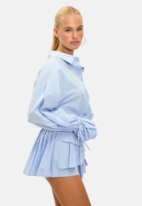 Robe chemise bleu clair avec des manches surdimensionnées, des poignets froncés et une jupe plissée à plusieurs couches. Fabriquée dans un tissu lisse avec un col ajusté.