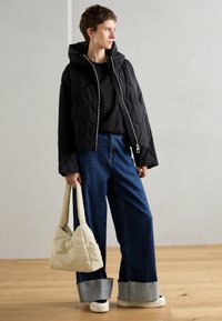 Veste noire matelassée avec capuche, associée à un jean bleu ample et un sac à main couleur crème. Baskets blanches et détails gris au niveau des poignets du jean.