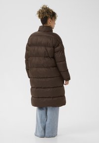 Hnedá prešívaná bunda s vysokým golierom, prešitými časťami a voľným strihom, kombinovaná s širokými džínsami v svetlom modrom denimovom materiáli.