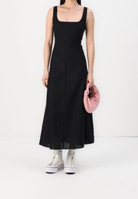 Svart ärmlös midiklänning med fyrkantig halsringning, tillverkad av lättviktig tyg, kombinerad med en rosa knuten clutch och vita sneakers.