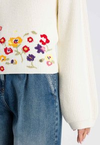 Maglione color crema con texture a coste e ricami floreali in rosso, giallo e viola, abbinato a jeans in denim azzurri.