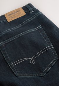 Tumed denimist valmistatud teksased, millel on nahkplekk, millel on kiri "DENIM SUPPLY." Taga taskul on märgatav valge tikand kõveras mustris.