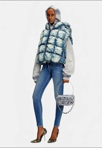 Γυναίκα με γκρι φούτερ, μπλε αδιάβροχη επένδυση tie-dye, στενά τζιν, παραλλαγής τακούνια, κρατώντας μεταλλική ασημί τσάντα χειρός με αλυσίδα.