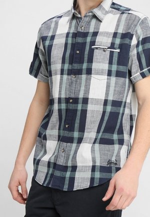 Man draagt een overhemd met korte mouwen, een geruit patroon in marineblauw, grijs, wit en groen, met een borstzakje voorzien van knoopdetail.