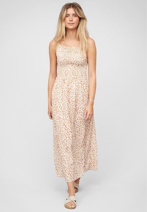 Jonge vrouw met lang blond haar, gekleed in een mouwloze, lichtroze maxi-jurk met bruine vlekken en beige sandalen, staand tegen een effen achtergrond.