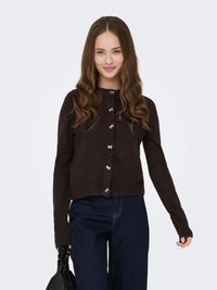 Braune gestrickte Strickjacke mit rundem Ausschnitt, ausgestattet mit goldfarbenen dekorativen Knöpfen, langen Ärmeln und einer kurzen Passform, kombiniert mit dunklen Jeans.