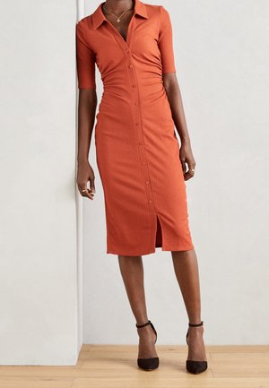 Gestreiftes, knopfmeshtes Kleid in verbranntem Orange, mit kurzen Ärmeln, einem Kragen und einem schulterhohen Schlitz. Gestylt mit schwarzen Absatzsandalen mit Knöchelriemen.