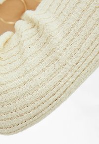 Chausson en tricot beige à texture, de forme incurvée, présentant un motif côtelé subtil et un matériau doux et duveteux. Sans accessoires ni détails visibles.