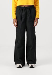 Pantalons cargo noirs avec une coupe ample, des plis, des poches latérales et une taille élastique. Portés avec un haut en polaire jaune court et des baskets claires.