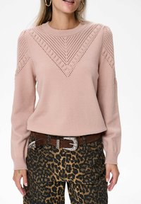 Kobieta w jasnoróżowym swetrze z dzianiny, z warkoczowymi i ażurowymi detalami, w połączeniu z spodniami w leopardzi wzór oraz brązowym paskiem z srebrną klamrą.