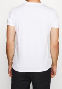 Bílý tričko s krátkým rukávem, kulatým výstřihem a rovný střih. Měkká látka s hladkou texturou, na spodním lemu je malá nášivka.