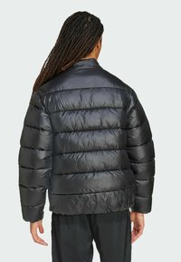 Henkilö, jolla on pitkät tummat hiukset, pukeutuneena mustaan paksuun takkiin, seisoo selin, vaaleanharmaata taustaa vasten.