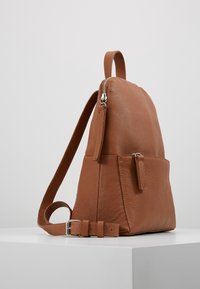 Sac à dos en cuir marron avec une finition texturée, fermeture éclair, bandoulière réglable et une poche avant avec un petit détail zippé.