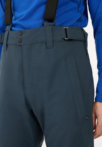 Donkergrijze technische broek met verstelbare bretels, twee grote voorknopjes en een zijzak, kenmerkt zich door een strakke, gladde stoftextuur.