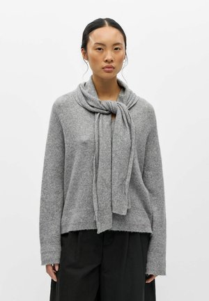 Jeune femme aux cheveux foncés porte un pull gris doux avec une écharpe assortie nouée autour du cou et un pantalon noir large sur un fond blanc uni.