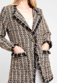 Tweedblazer i svart, beige och vitt med ett strukturerat mönster, fransade kanter och framfickor. Har en enda knäppning med mässingsknapp.