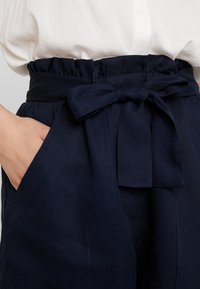 Námořnické látkové kalhoty se shromážděným pasem a předním vázáním. Mají boční kapsy a texturovaný povrch.