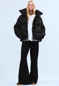Čierna puff jacket s vysokým golierom, skombinovaná s bielym topom a širokými čiernymi nohavicami. Modelka má dlhé vlasy a priehľadné okuliare.