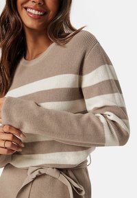 Maglione a righe beige e crema con un filo lavorato a maglia. Presenta un collo a giro e maniche relax raccolte ai polsini.