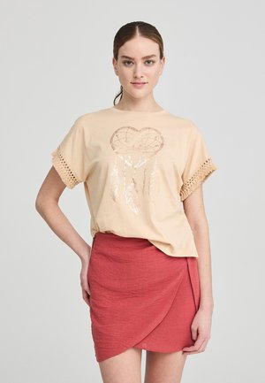 T-shirt beige con design a forma di cuore di un dreamcatcher e accenti di piume, con maniche in crochet con frange. Abbinato a una gonna rossa a portafoglio.