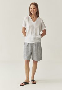 Top alb cu mânecă scurtă și decolteu în V, dintr-un material texturat, asociat cu pantaloni scurți negri și albi, cu dungi, până la genunchi. Sandale negre cu talpă plata.