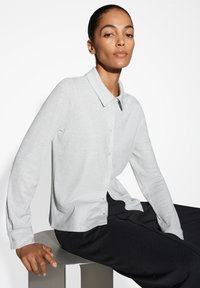 Camicia a maniche lunghe grigia chiara con bottoni e colletto, realizzata in un tessuto testurizzato e brillante. Abbinata a pantaloni neri, modello seduto su uno sgabello.