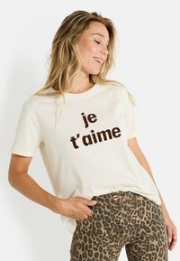 Mujer sonriente con camisa crema con texto de leopardo "je t'aime", lleva pantalones de leopardo, mano en el bolsillo, mirando hacia un lado.