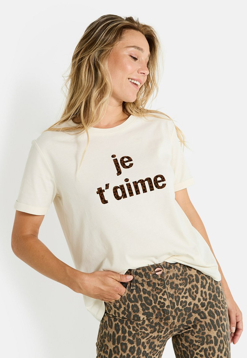 Mujer sonriente con camisa crema con texto de leopardo "je t'aime", lleva pantalones de leopardo, mano en el bolsillo, mirando hacia un lado.