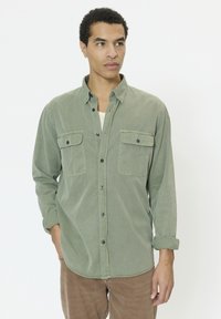 Noor mees, kes kannab helerohelist pika varrukaga nööbitavat särki kahe rinnataskuga ja pruune pükse, vaadates kõrvale.