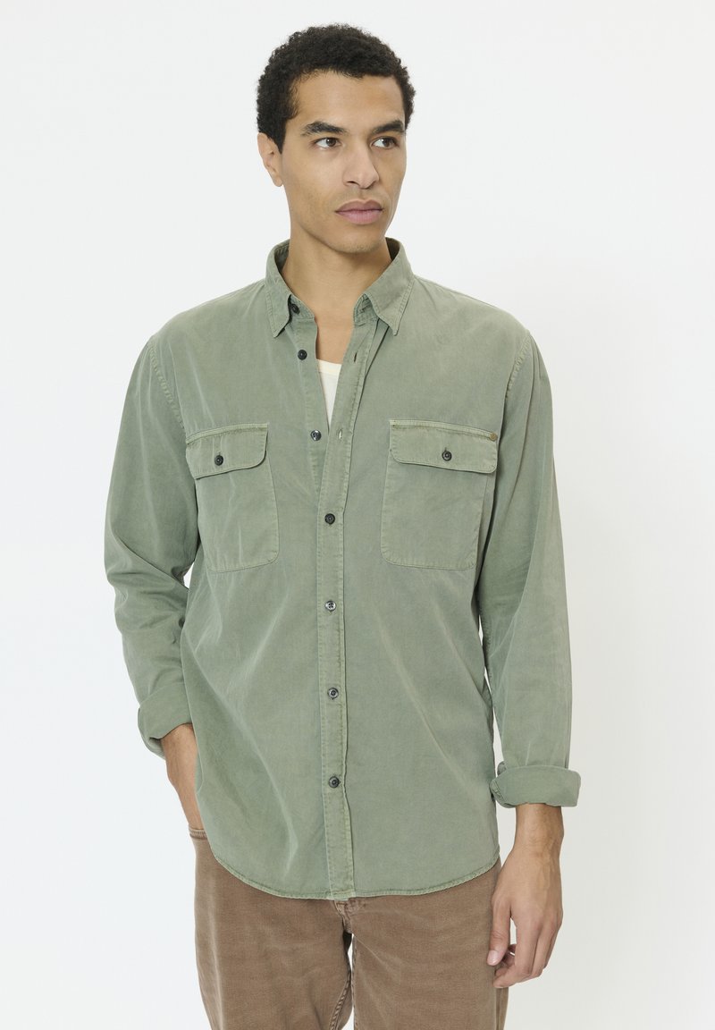 Noor mees, kes kannab helerohelist pika varrukaga nööbitavat särki kahe rinnataskuga ja pruune pükse, vaadates kõrvale.