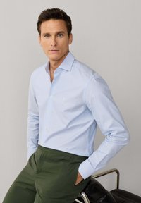 Homme aux cheveux bruns courts portant une chemise à boutons bleu clair et un pantalon vert olive, debout les mains dans les poches devant un fond uni.