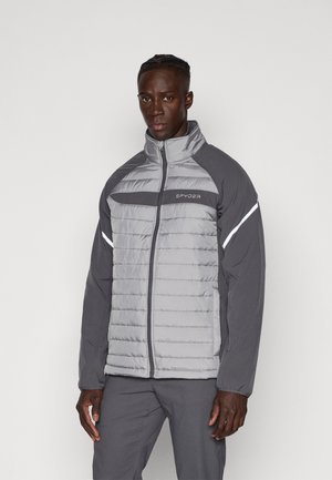 Uomo che indossa una giacca trapuntata grigia con zip, maniche grigio scuro e strisce riflettenti sulle braccia, in piedi davanti a uno sfondo chiaro e uniforme.