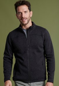 Giacca in pile nera con colletto alto, zip frontale completa e tasche laterali con zip. Presenta una superficie testurizzata e un design slim.