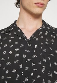 Maglietta nera a maniche corte con motivi grafici bianchi che includono soli, arcobaleni e motivi tropicali. Presenta un colletto a camicia e una patta con bottoni.