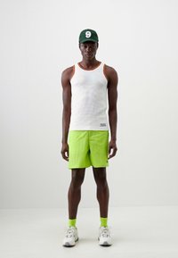 Hvit ribbet tanktopp med oransje detaljer, lys grønne shorts, grønn caps og hvite joggesko med aksenter. Modell som står mot en ensfarget bakgrunn.