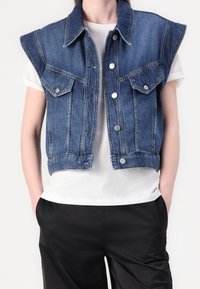 Denim bezrukává vesta ve středně modrém odstínu s dvěma předními kapsami, patentními knoflíky a klasickým límcem. Nositá přes bílé tričko.