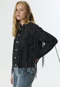 Cardigan tejido en blanco y negro con botones en la parte frontal, detalles de flecos con acentos metálicos y mangas largas. Combinado con unos vaqueros de color azul claro.