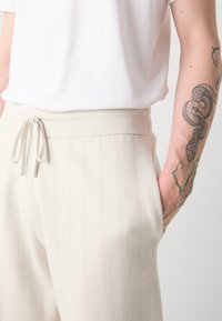Pantalones de color crema con rayas verticales de textura, cinturilla elástica con cordón ajustable y bolsillos laterales. Acabado de tela suave.