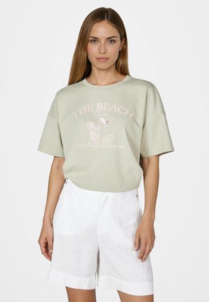 Woman wearing a light green graphic t-shirt with "The Beach" text and white high-waisted shorts, standing against a plain white background.