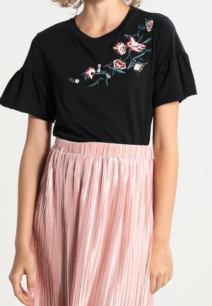 Czarny krótki top z kwiatowym haftem w połączeniu z jasnoróżową plisowaną spódnicą z wysokim stanem na stojącej osobie.