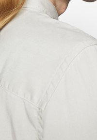Camicetta in cotone beige chiaro con colletto rialzato, cuciture visibili e una finitura testurizzata. Il tessuto appare morbido e resistente.