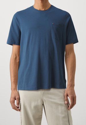 Mand iført en kortærmet blå t-shirt med rund hals og brystlomme samt beige bukser mod en ensfarvet baggrund.