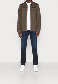 Brun lynlåstrøje med to brystlommer, lagdelt over en hvid t-shirt. Kombineret med mørkeblå jeans og hvide sneakers.