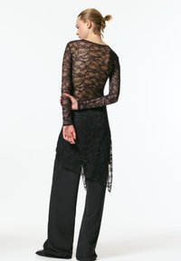 Bluza neagră cu mâneci lungi din dantelă cu model floral, purtată peste o fustă neagră, asimetrică și mulată, și pantaloni negri largi. Design transparent și texturat.