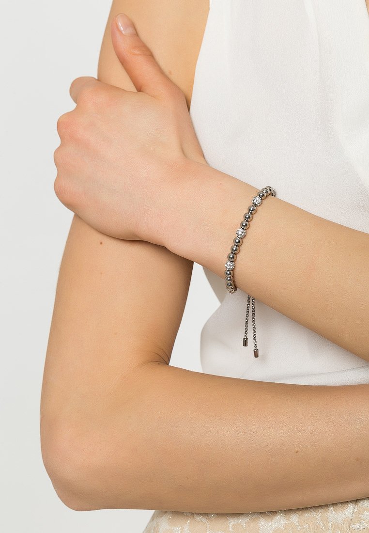 Srebrna bransoletka z koralikami, w której przeplatają się gładkie i fakturowane kuleczki, z regulowanym zapięciem i dwoma metalowymi końcówkami, noszona na nadgarstku.