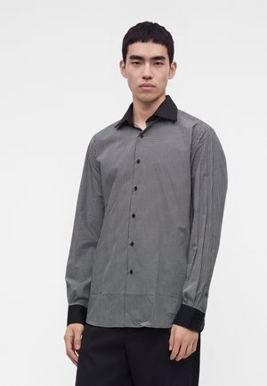 Jeune homme portant une chemise à manches longues noire et blanche à motif avec col et poignets noirs, se tenant devant un fond blanc uni.