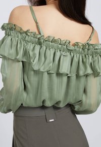 Top off-the-shoulder di colore verde chiaro, realizzato in tessuto trasparente con righe verticali, dettagli arricciati e bretelle regolabili.