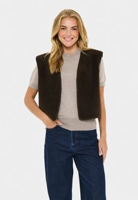 Mujer sonriente con cabello rubio, vistiendo un suéter beige de manga corta, un chaleco sin mangas marrón oscuro y jeans azul oscuro, de pie con una mano en el bolsillo.