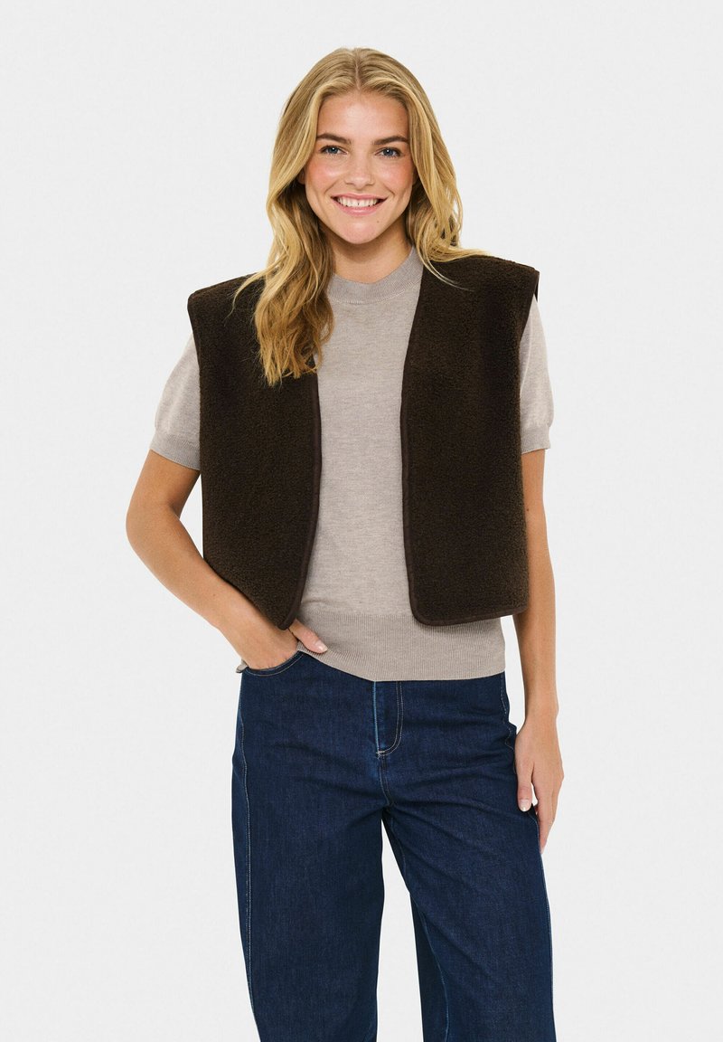 Mujer sonriente con cabello rubio, vistiendo un suéter beige de manga corta, un chaleco sin mangas marrón oscuro y jeans azul oscuro, de pie con una mano en el bolsillo.