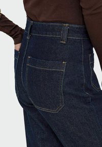 Jeans en denim foncé taille haute avec une texture lisse, des coutures jaunes contrastantes et deux poches arrière.